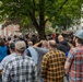 A large outdoor crowd of predominantly adult males has gathered in what appears to be a public square or plaza near brick institutional buildings. One individual in the foreground right is wearing a white hard hat marked with the number '712', suggesting union or trade worker affiliation. The crowd appears to be attending a rally, demonstration, or public gathering, with people facing toward a central focal point obscured by trees.
