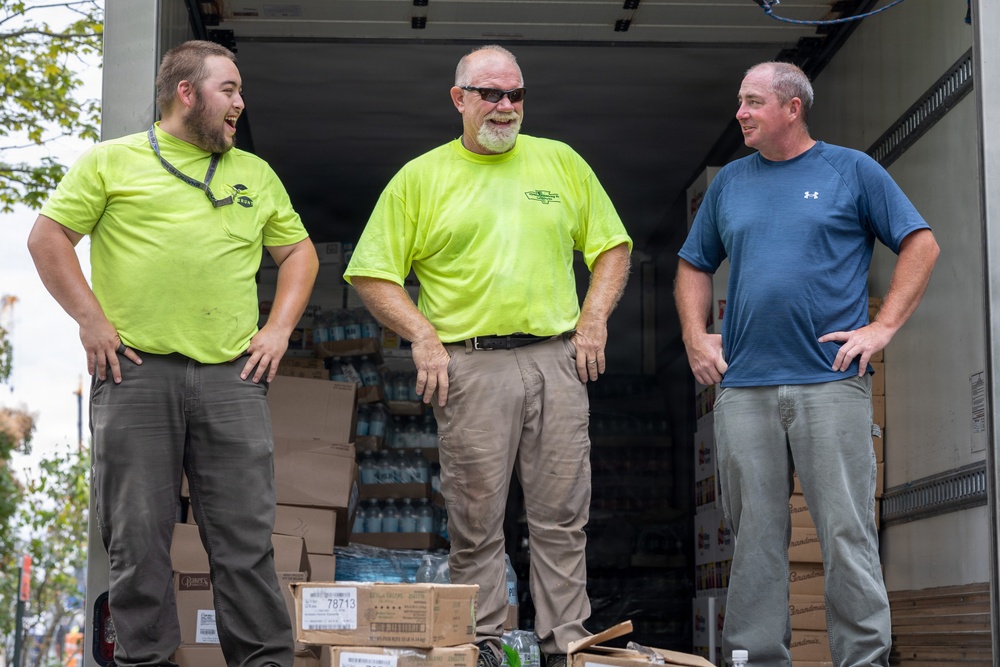 Three men are standing at the open rear of a delivery truck loaded with boxes and cases of bottled water. Two of the men are wearing matching neon yellow/lime green shirts, suggesting they are coworkers or part of the same organization. The group appears to be in good spirits, laughing and conversing during what appears to be a supply delivery or distribution operation.