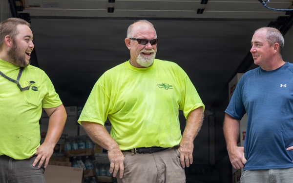 Three men are standing at the open rear of a delivery truck loaded with boxes and cases of bottled water. Two of the men are wearing matching neon yellow/lime green shirts, suggesting they are coworkers or part of the same organization. The group appears to be in good spirits, laughing and conversing during what appears to be a supply delivery or distribution operation.
