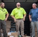 Three men are standing at the open rear of a delivery truck loaded with boxes and cases of bottled water. Two of the men are wearing matching neon yellow/lime green shirts, suggesting they are coworkers or part of the same organization. The group appears to be in good spirits, laughing and conversing during what appears to be a supply delivery or distribution operation.