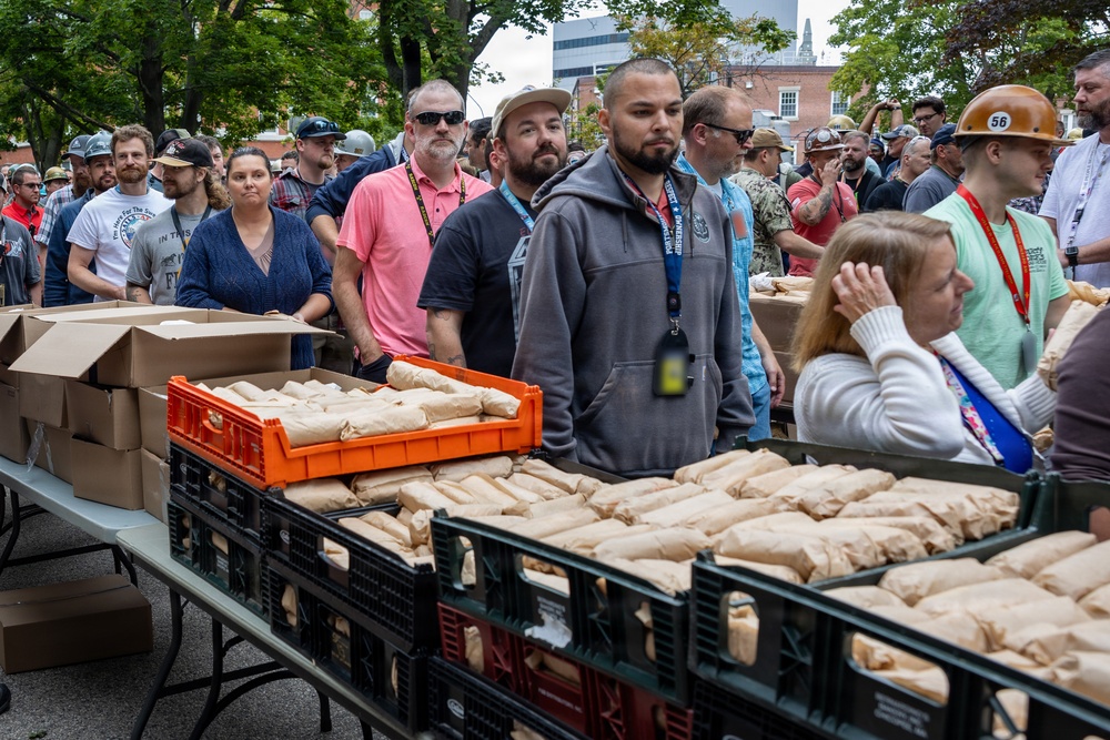 A large crowd of people is gathered outdoors around long folding tables loaded with crates and boxes containing paper-wrapped items, likely food packages or bag lunches. The setting appears to be an organized distribution event, possibly a community meal service, charity food giveaway, or worker appreciation event. Several individuals are wearing hard hats, lanyards, and ID badges, suggesting an industrial or organized event context.