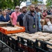 A large crowd of people is gathered outdoors around long folding tables loaded with crates and boxes containing paper-wrapped items, likely food packages or bag lunches. The setting appears to be an organized distribution event, possibly a community meal service, charity food giveaway, or worker appreciation event. Several individuals are wearing hard hats, lanyards, and ID badges, suggesting an industrial or organized event context.