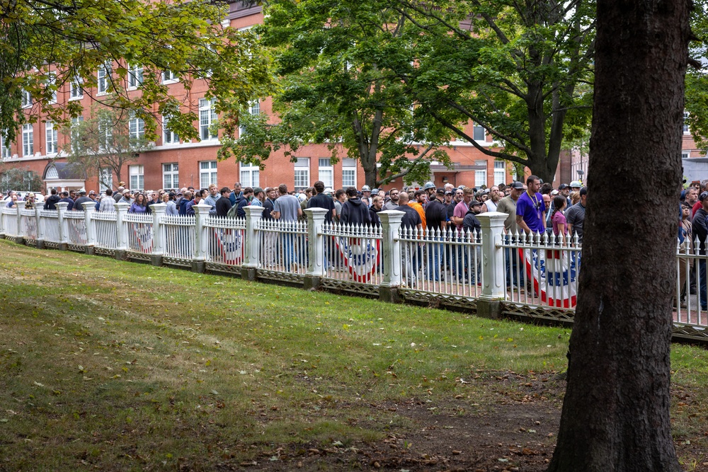 A large crowd of people is gathered along a white decorative iron fence in what appears to be a public park or common area. Red, white, and blue bunting or banners are draped along the fence, suggesting a patriotic or civic event. A large red brick institutional building is visible in the background, surrounded by mature green trees.