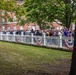 A large crowd of people is gathered along a white decorative iron fence in what appears to be a public park or common area. Red, white, and blue bunting or banners are draped along the fence, suggesting a patriotic or civic event. A large red brick institutional building is visible in the background, surrounded by mature green trees.