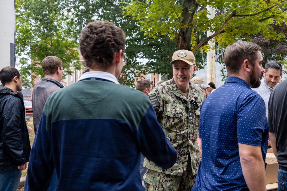 A person in U.S. Navy digital camouflage (NWU Type III) uniform and tan cap is engaged in conversation with civilians at an outdoor gathering. The name tape on the uniform reads 'NICE'. The setting appears to be a community or campus event with multiple people mingling near trees and brick buildings.