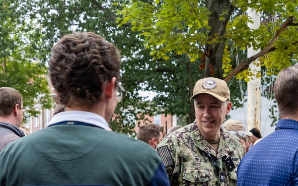 A person in U.S. Navy digital camouflage (NWU Type III) uniform and tan cap is engaged in conversation with civilians at an outdoor gathering. The name tape on the uniform reads 'NICE'. The setting appears to be a community or campus event with multiple people mingling near trees and brick buildings.