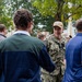 A person in U.S. Navy digital camouflage (NWU Type III) uniform and tan cap is engaged in conversation with civilians at an outdoor gathering. The name tape on the uniform reads 'NICE'. The setting appears to be a community or campus event with multiple people mingling near trees and brick buildings.