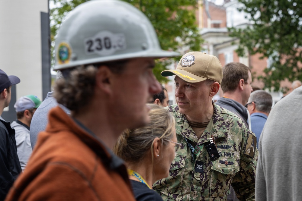 A U.S. Navy officer in NWU (Navy Working Uniform) camouflage is engaged in conversation with a group of civilians, one of whom is wearing a hard hat numbered '230'. The naval officer appears to hold the rank of Captain (O-6), indicated by the eagle insignia visible on the collar. The setting appears to be an outdoor urban environment, possibly a construction site visit or official event.