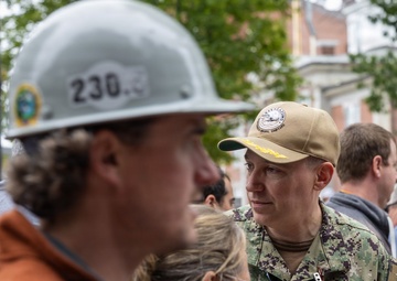 A U.S. Navy officer in NWU (Navy Working Uniform) camouflage is engaged in conversation with a group of civilians, one of whom is wearing a hard hat numbered '230'. The naval officer appears to hold the rank of Captain (O-6), indicated by the eagle insignia visible on the collar. The setting appears to be an outdoor urban environment, possibly a construction site visit or official event.