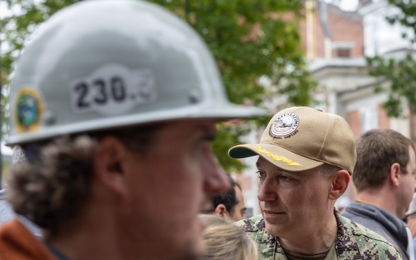 A U.S. Navy officer in NWU (Navy Working Uniform) camouflage is engaged in conversation with a group of civilians, one of whom is wearing a hard hat numbered '230'. The naval officer appears to hold the rank of Captain (O-6), indicated by the eagle insignia visible on the collar. The setting appears to be an outdoor urban environment, possibly a construction site visit or official event.