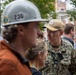 A U.S. Navy officer in NWU (Navy Working Uniform) camouflage is engaged in conversation with a group of civilians, one of whom is wearing a hard hat numbered '230'. The naval officer appears to hold the rank of Captain (O-6), indicated by the eagle insignia visible on the collar. The setting appears to be an outdoor urban environment, possibly a construction site visit or official event.