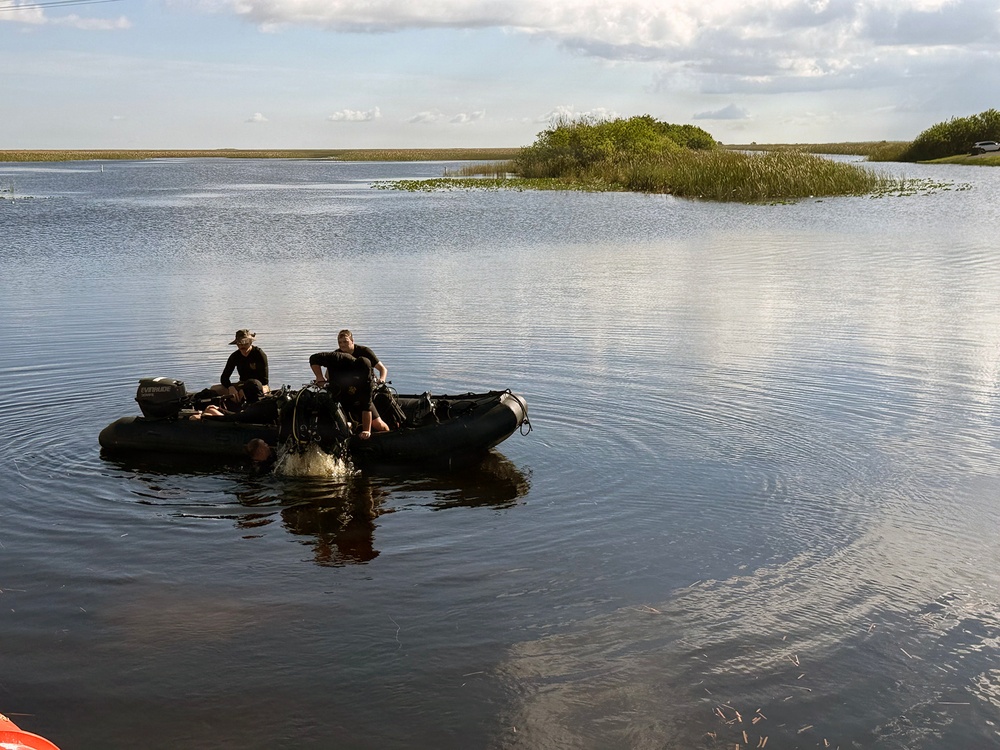 Two individuals dressed in black are aboard a dark inflatable rubber boat (likely a Zodiac-type craft) on a calm, shallow waterway surrounded by marsh vegetation. The boat is equipped with an outboard motor and what appears to be diving or tactical gear. The setting resembles a wetland or estuary environment, possibly the Florida Everglades or similar subtropical marsh.
