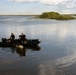 Two individuals dressed in black are aboard a dark inflatable rubber boat (likely a Zodiac-type craft) on a calm, shallow waterway surrounded by marsh vegetation. The boat is equipped with an outboard motor and what appears to be diving or tactical gear. The setting resembles a wetland or estuary environment, possibly the Florida Everglades or similar subtropical marsh.