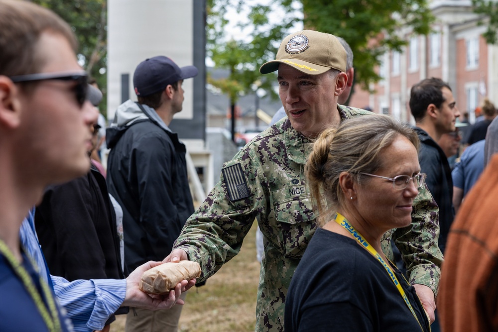 A U.S. Navy service member in NWU Type III camouflage uniform with the name tape 'NICE' is shaking hands with a civilian at what appears to be an outdoor public event. The setting includes brick buildings and trees in the background, suggesting a military installation or academy campus. Several other civilians and personnel are visible in the crowd.