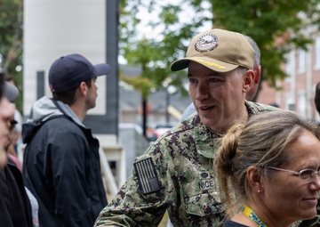 A U.S. Navy service member in NWU Type III camouflage uniform with the name tape 'NICE' is shaking hands with a civilian at what appears to be an outdoor public event. The setting includes brick buildings and trees in the background, suggesting a military installation or academy campus. Several other civilians and personnel are visible in the crowd.