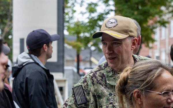 A U.S. Navy service member in NWU Type III camouflage uniform with the name tape 'NICE' is shaking hands with a civilian at what appears to be an outdoor public event. The setting includes brick buildings and trees in the background, suggesting a military installation or academy campus. Several other civilians and personnel are visible in the crowd.
