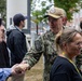 A U.S. Navy service member in NWU Type III camouflage uniform with the name tape 'NICE' is shaking hands with a civilian at what appears to be an outdoor public event. The setting includes brick buildings and trees in the background, suggesting a military installation or academy campus. Several other civilians and personnel are visible in the crowd.