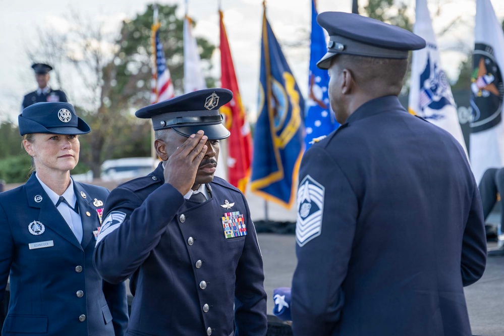 A formal U.S. Space Force or Air Force military ceremony is taking place outdoors, featuring personnel in dress uniforms. A service member in the center is rendering a salute to a senior enlisted member facing away from the camera, while a female officer stands to the left observing. Multiple military branch flags are displayed in the background.