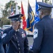 A formal U.S. Space Force or Air Force military ceremony is taking place outdoors, featuring personnel in dress uniforms. A service member in the center is rendering a salute to a senior enlisted member facing away from the camera, while a female officer stands to the left observing. Multiple military branch flags are displayed in the background.