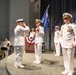 A formal U.S. Navy change of command ceremony is taking place on a stage. Officers in dress white uniforms are participating in the transfer of a unit guidon (flag), with one officer saluting while another holds the blue flag. A military band is visible in the background left, and an American flag is displayed on the right side of the stage.