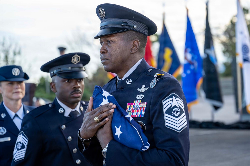 A United States Air Force senior non-commissioned officer, identifiable by Chief Master Sergeant rank insignia, holds a folded American flag during what appears to be a formal military ceremony. Multiple service members in dress blue uniforms are present, with a color guard and various military flags visible in the background. The solemn expressions and ceremonial posture suggest this is a retirement, memorial, or honors ceremony.
