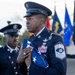 A United States Air Force senior non-commissioned officer, identifiable by Chief Master Sergeant rank insignia, holds a folded American flag during what appears to be a formal military ceremony. Multiple service members in dress blue uniforms are present, with a color guard and various military flags visible in the background. The solemn expressions and ceremonial posture suggest this is a retirement, memorial, or honors ceremony.