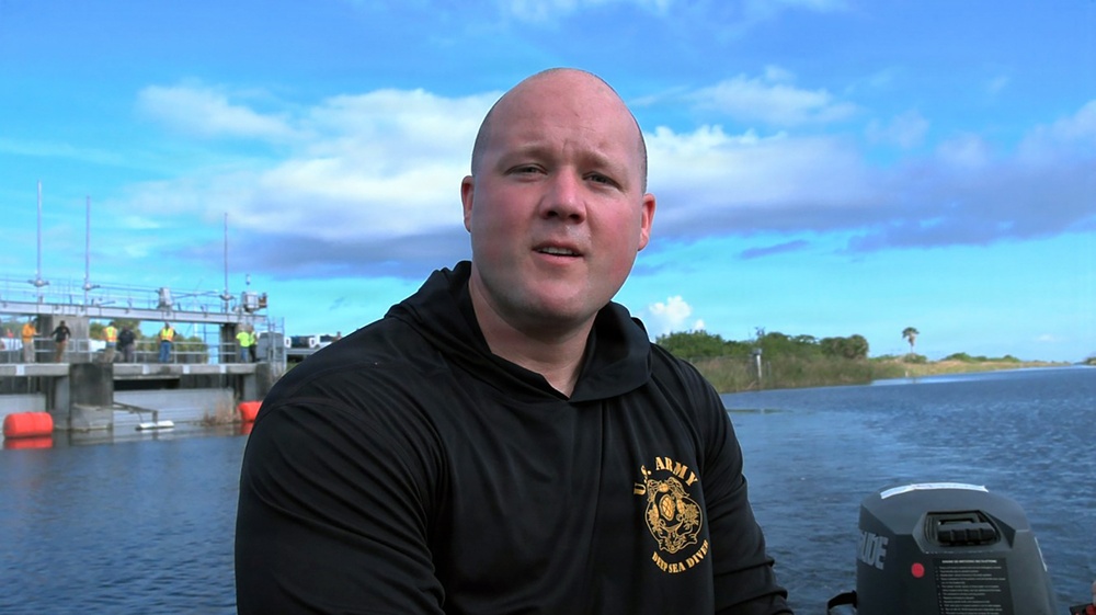 A bald male individual wearing a black 'U.S. Army Deep Sea Diver' hoodie is seated on or near a small boat on a waterway. He appears to be speaking, possibly during an interview or briefing. In the background, a water control structure or dam is visible with several people standing on it.