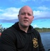 A bald male individual wearing a black 'U.S. Army Deep Sea Diver' hoodie is seated on or near a small boat on a waterway. He appears to be speaking, possibly during an interview or briefing. In the background, a water control structure or dam is visible with several people standing on it.