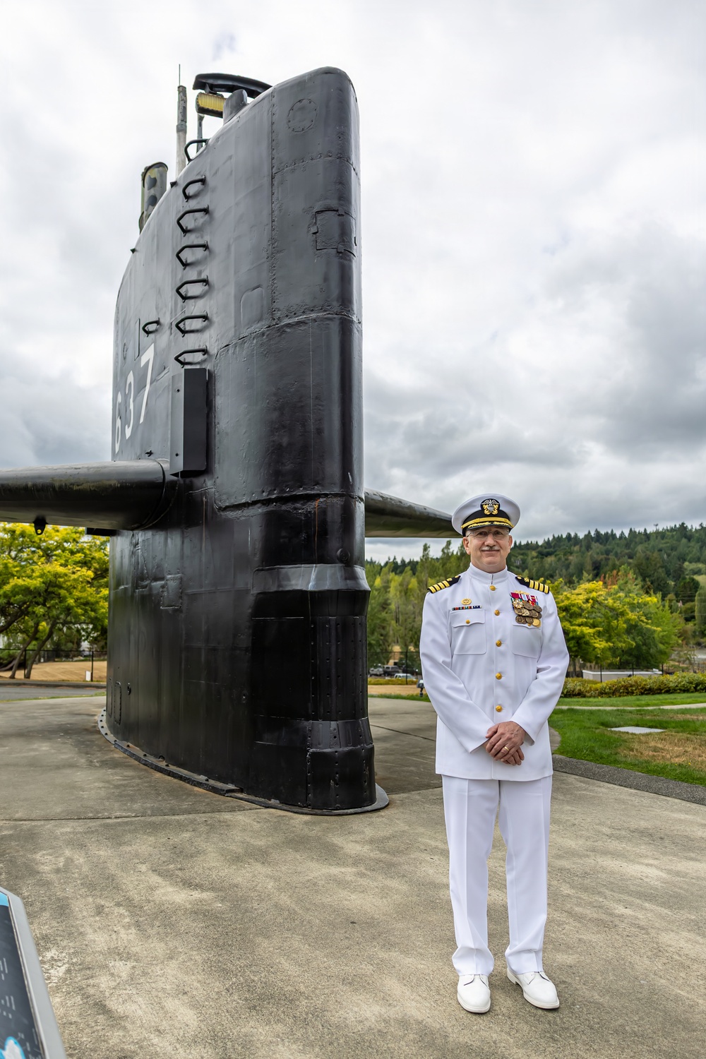 A U.S. Navy officer in white dress uniform stands beside the sail (conning tower) of a submarine on static display, marked with the hull number 637. The setting appears to be an outdoor naval museum or memorial, surrounded by trees and manicured grounds. The officer is posing formally with hands clasped in front.