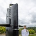 A U.S. Navy officer in white dress uniform stands beside the sail (conning tower) of a submarine on static display, marked with the hull number 637. The setting appears to be an outdoor naval museum or memorial, surrounded by trees and manicured grounds. The officer is posing formally with hands clasped in front.