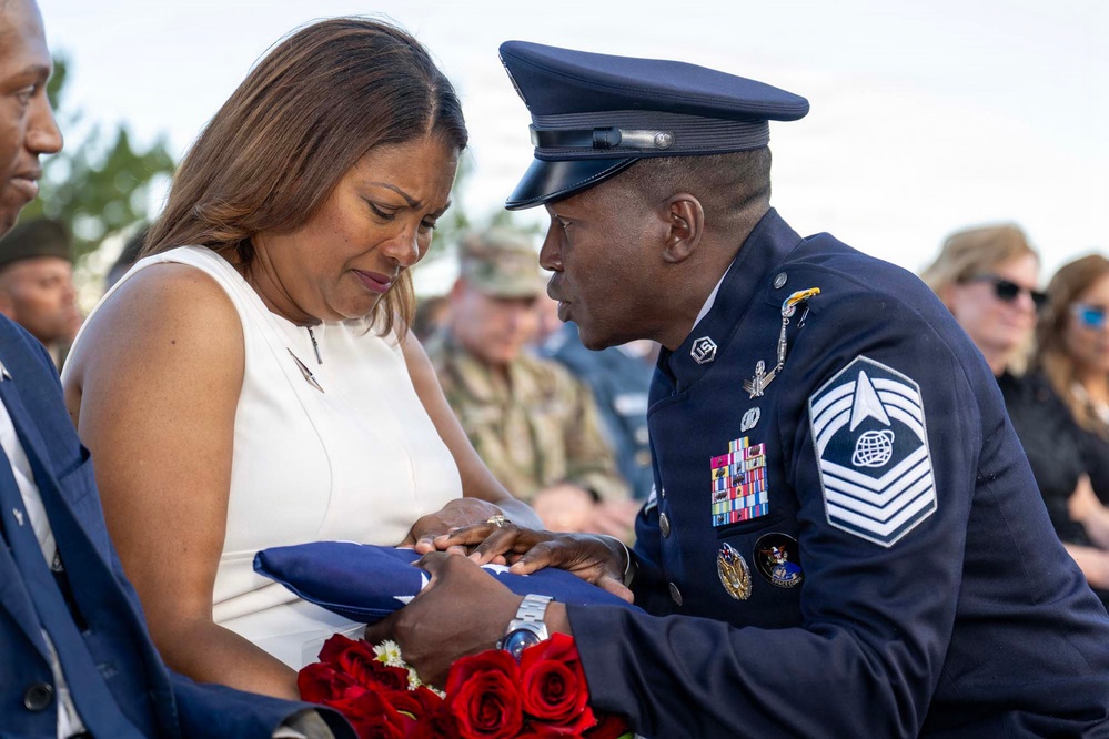 A U.S. Air Force senior non-commissioned officer in dress blues is presenting a folded American flag to a grieving woman in a white dress during what appears to be a military funeral or memorial service. The woman is visibly emotional, holding both the flag and red roses. The scene is surrounded by other attendees, some in military uniform, consistent with a formal military burial ceremony.