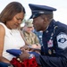A U.S. Air Force senior non-commissioned officer in dress blues is presenting a folded American flag to a grieving woman in a white dress during what appears to be a military funeral or memorial service. The woman is visibly emotional, holding both the flag and red roses. The scene is surrounded by other attendees, some in military uniform, consistent with a formal military burial ceremony.