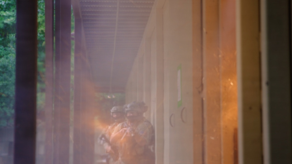 The image shows what appears to be military or tactical personnel positioned along a corridor or building exterior, partially obscured by smoke or haze with an orange tint. At least two figures wearing helmets and tactical gear are visible in the middle distance. The scene suggests a training exercise or tactical operation in a structured environment.
