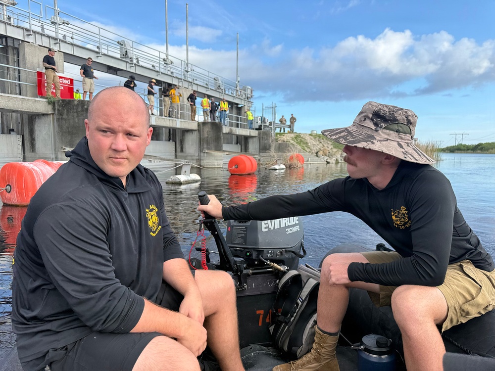 Two men in matching black long-sleeve shirts are seated in a small inflatable boat equipped with an Evinrude 30MFE outboard motor on a body of water near a concrete flood control structure or dam. Orange buoys are visible in the water, and a group of people wearing safety vests and civilian clothing are gathered on the structure in the background. The scene appears to be a water management or emergency response operation.