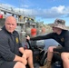 Two men in matching black long-sleeve shirts are seated in a small inflatable boat equipped with an Evinrude 30MFE outboard motor on a body of water near a concrete flood control structure or dam. Orange buoys are visible in the water, and a group of people wearing safety vests and civilian clothing are gathered on the structure in the background. The scene appears to be a water management or emergency response operation.