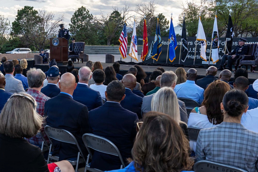 An outdoor military ceremony is taking place with a uniformed officer speaking at a podium on a stage. A large audience seated in folding chairs faces the stage, which displays multiple military and national flags. A banner in the background references 'Headquarters Combat Forces' and 'U.S. Space Force,' suggesting this is an official Space Force command ceremony.
