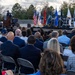 An outdoor military ceremony is taking place with a uniformed officer speaking at a podium on a stage. A large audience seated in folding chairs faces the stage, which displays multiple military and national flags. A banner in the background references 'Headquarters Combat Forces' and 'U.S. Space Force,' suggesting this is an official Space Force command ceremony.