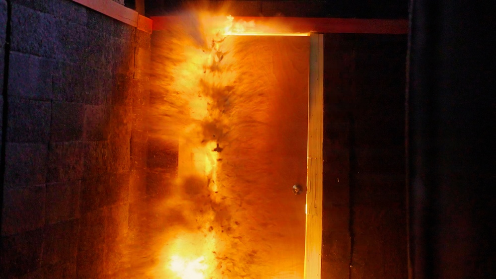 The image shows an open doorway with intense fire and flames erupting from within a room, illuminating the surrounding dark walls with bright orange and yellow light. The fire appears to be actively burning inside the room and is visible through the open door frame. This appears to be a fire safety demonstration, controlled burn, or structural fire scenario.