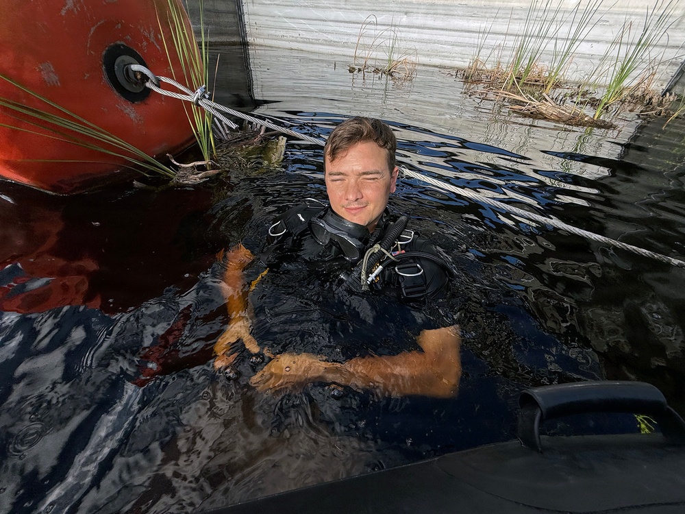 A male diver in full scuba gear is submerged in dark, murky water near what appears to be a boat hull painted red and a metal structure. He is holding a wet, orange-colored cat or small animal above the water surface. The setting appears to be a flooded or waterlogged area with vegetation visible in the background.