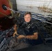 A male diver in full scuba gear is submerged in dark, murky water near what appears to be a boat hull painted red and a metal structure. He is holding a wet, orange-colored cat or small animal above the water surface. The setting appears to be a flooded or waterlogged area with vegetation visible in the background.