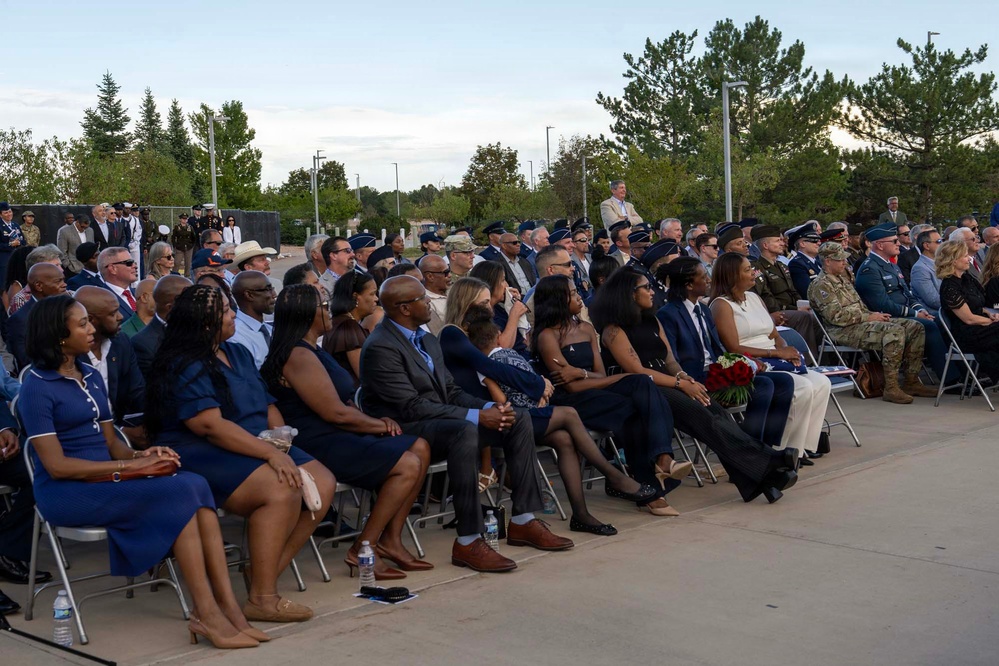 A large outdoor ceremony is taking place with a diverse audience seated in folding chairs on a paved surface. The crowd includes both civilians in formal attire and military personnel in various uniforms, suggesting an official military or government event. Trees and light poles are visible in the background under a partly cloudy sky.