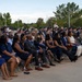 A large outdoor ceremony is taking place with a diverse audience seated in folding chairs on a paved surface. The crowd includes both civilians in formal attire and military personnel in various uniforms, suggesting an official military or government event. Trees and light poles are visible in the background under a partly cloudy sky.