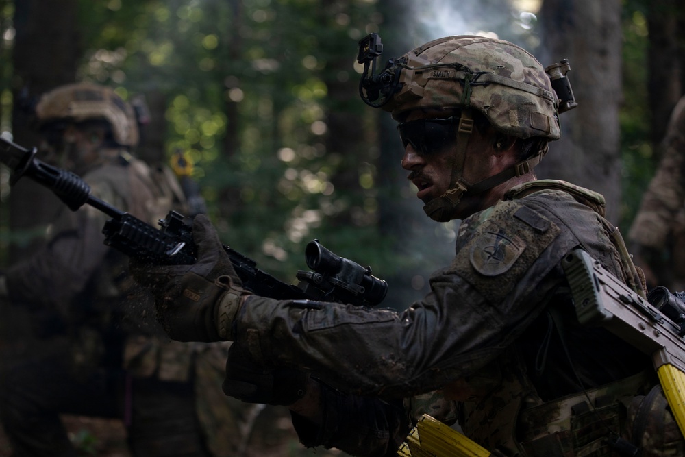 Two soldiers in full combat gear are operating in a forested environment. The foreground soldier is clearly visible wearing Operational Camouflage Pattern (OCP) uniform with a helmet-mounted device, tactical eyewear, and holding a rifle with an optic attachment. A second soldier is visible in the background, also armed and in similar attire.