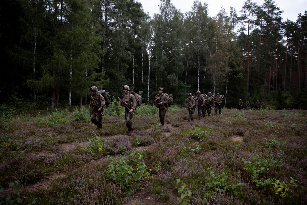 A column of soldiers in camouflage uniforms and full combat gear marches through a clearing with low-growing heather vegetation at the edge of a mixed forest. The soldiers are carrying rifles and large rucksacks, moving in a single-file or loose column formation. The setting and vegetation suggest a Northern or Central European environment, consistent with a military exercise or patrol.