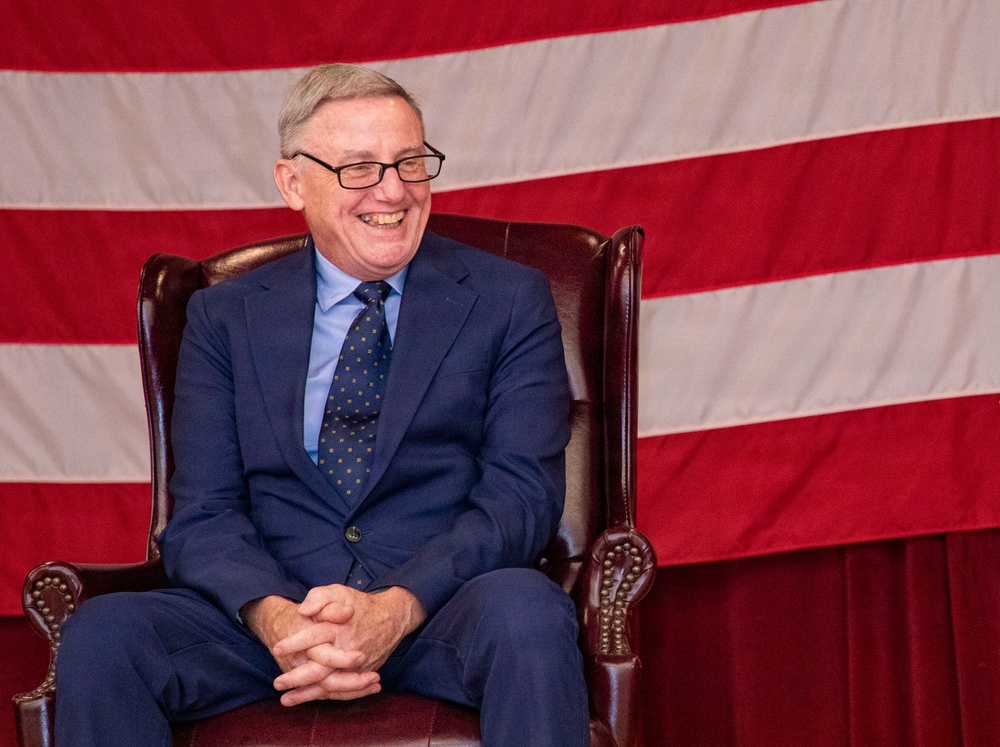 An older man wearing a navy blue suit and patterned tie is seated in a dark leather wingback chair, smiling broadly. He is positioned in front of a large American flag that serves as a backdrop. The setting appears formal, possibly a political or civic event.