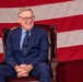 An older man wearing a navy blue suit and patterned tie is seated in a dark leather wingback chair, smiling broadly. He is positioned in front of a large American flag that serves as a backdrop. The setting appears formal, possibly a political or civic event.