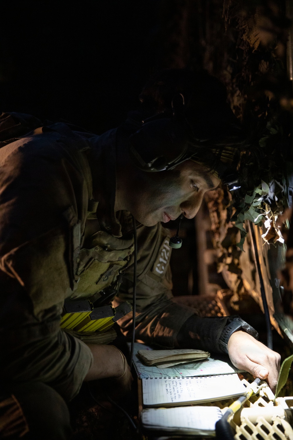 A military personnel is working in a low-light, concealed position, reviewing documents including what appears to be a map and a written notebook. The individual is equipped with tactical gear including a headlamp, body armor, and a radio or communication device. The setting appears to be a field position with camouflage netting visible in the background.
