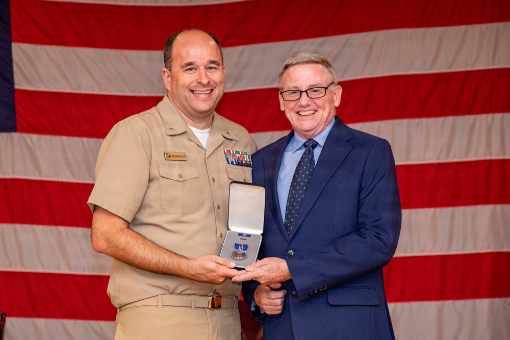 A U.S. Navy officer in Service Khaki uniform is posing with a civilian man in a blue suit, both holding an open medal presentation case. The ceremony appears to be a formal military award presentation taking place in front of a large American flag backdrop. The naval officer's name tag reads 'ROMANECHT' and he is wearing multiple service ribbons on his uniform.