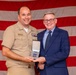 A U.S. Navy officer in Service Khaki uniform is posing with a civilian man in a blue suit, both holding an open medal presentation case. The ceremony appears to be a formal military award presentation taking place in front of a large American flag backdrop. The naval officer's name tag reads 'ROMANECHT' and he is wearing multiple service ribbons on his uniform.