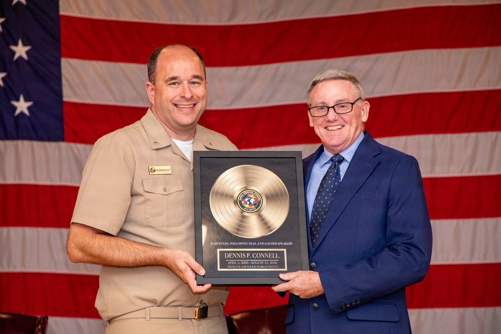 A U.S. Navy servicemember in tan/khaki uniform and a civilian man in a navy blue suit are jointly holding a commemorative gold record plaque in front of an American flag. The plaque honors Dennis P. Connell for his service from April 5, 2005 to August 31, 2026, with the inscription 'Fair Winds, Following Seas, and Louder Speakers.' This appears to be a military retirement or service recognition ceremony.